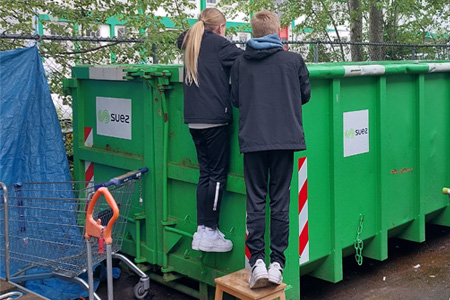 Leerlingen staan bij de rand van een grote groene container. Ze zijn op zoek naar bruikbare onderdelen om iets nieuws van te maken.
